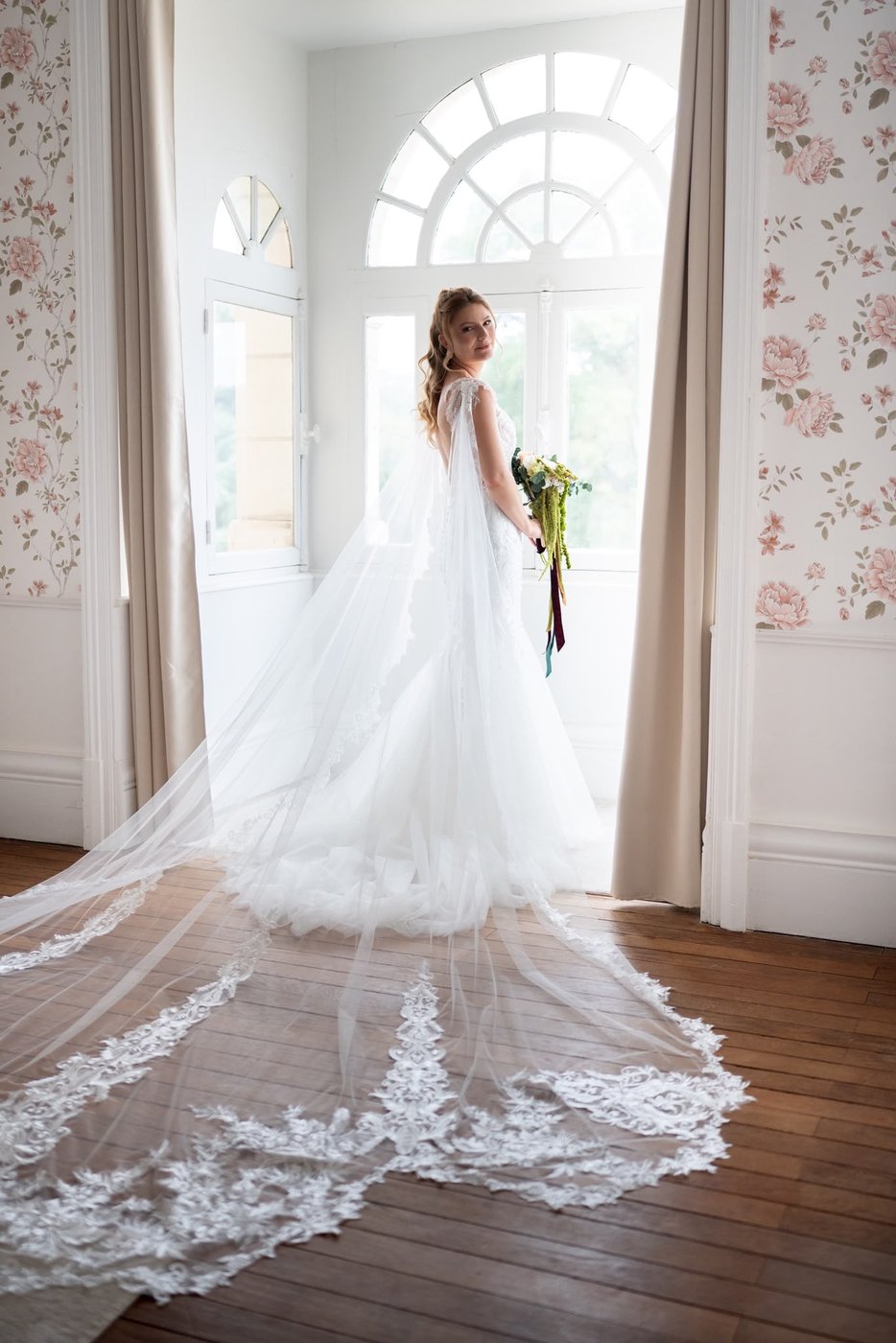 Mariée avec un long voile en dentelle devant une fenêtre de château